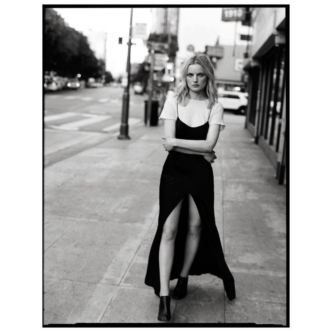 Full-length black and white shot of a woman in a black dress and white t-shirt standing on a city sidewalk with buildings in the background.