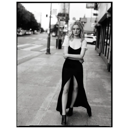 Full-length black and white shot of a woman in a black dress and white t-shirt standing on a city sidewalk with buildings in the background.