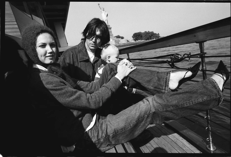 Black and white photograph of a family on a boat, featuring a woman with a knit hood, a man, and a baby, with the Statue of Liberty in the background.
