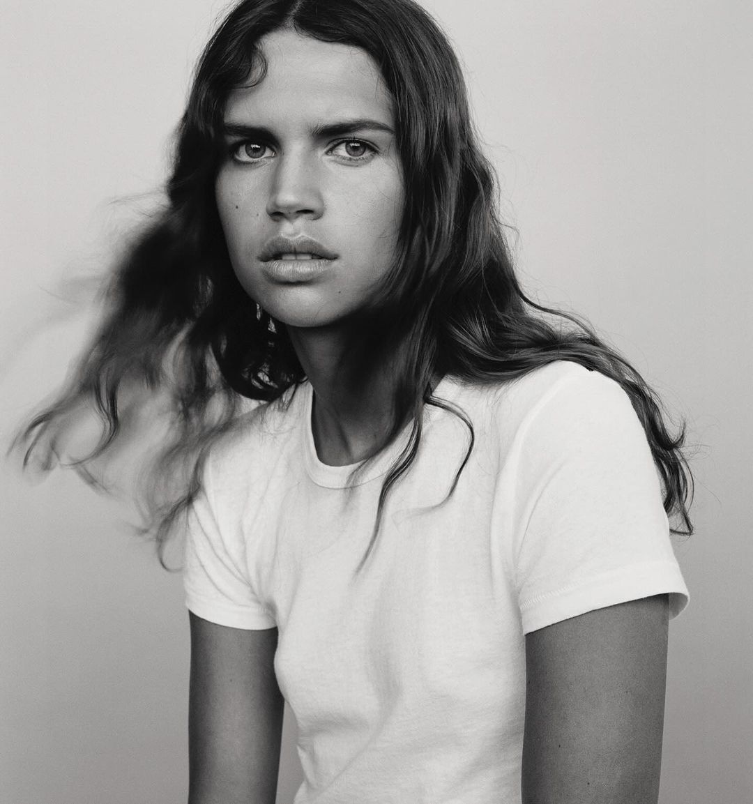 Portrait of a young woman with long dark wavy hair wearing a white t-shirt against a plain light grey backdrop.