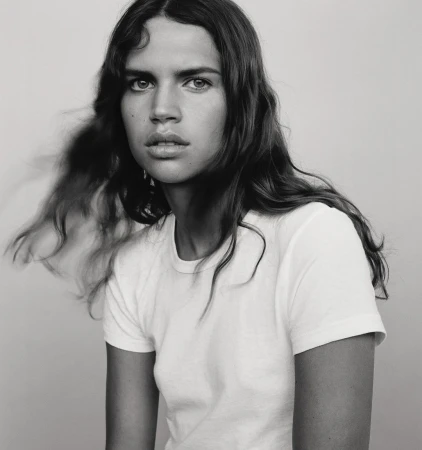 Portrait of a young woman with long dark wavy hair wearing a white t-shirt against a plain light grey backdrop.