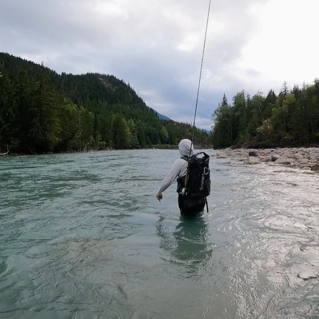 Fly fisherman wading in a glacial river surrounded by mountains and forest, wearing a fishing backpack, sport and outdoor activities.