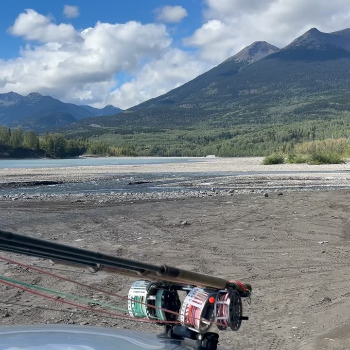 Fly fishing rods are mounted on a vehicle with a wide shot of a silty river, mountains, and cloudy skies in the background.