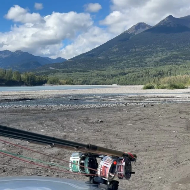 Fly fishing rods are mounted on a vehicle with a wide shot of a silty river, mountains, and cloudy skies in the background.
