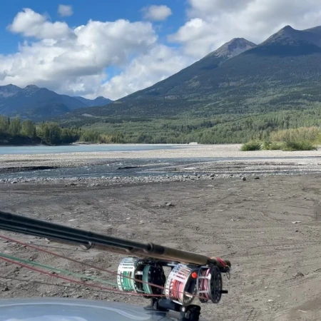 Fly fishing rods are mounted on a vehicle with a wide shot of a silty river, mountains, and cloudy skies in the background.