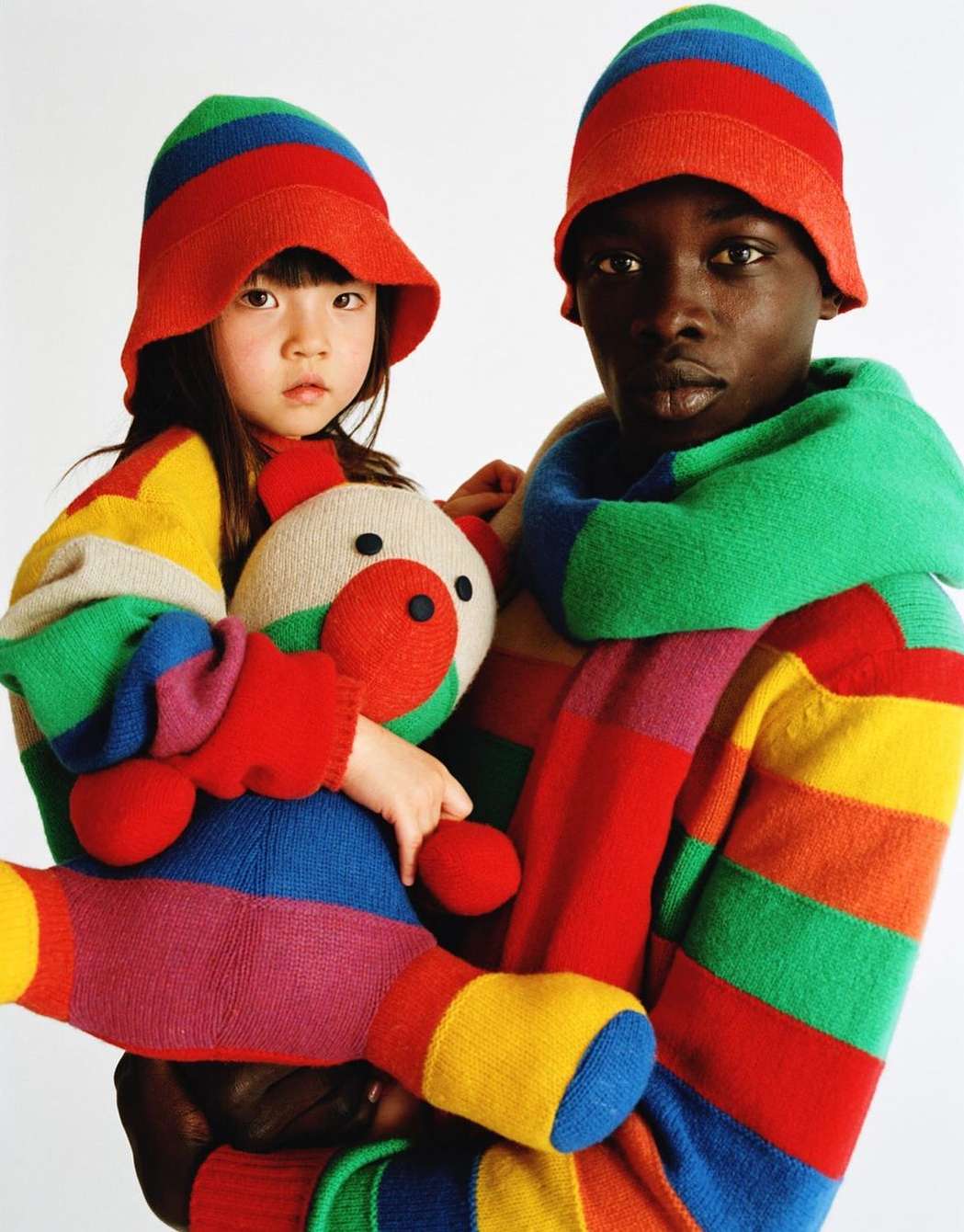 Two people, an adult and child, are wearing matching striped hats and outfits in a studio setting. The child holds a stuffed bear toy.