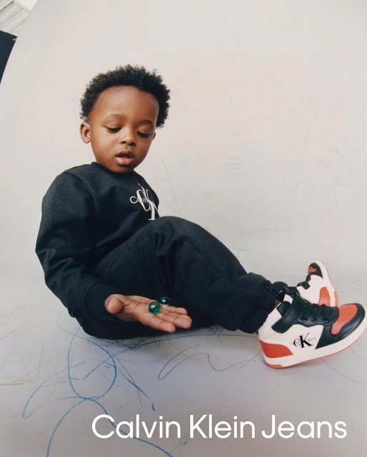 Child model sits with Calvin Klein Jeans branded shoes, holding green marbles in a light studio setting, showcasing a youth fashion brand.