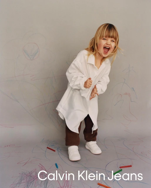 A happy toddler with blonde hair wears an oversized white top, brown pants, and white sneakers while standing in front of a crayon-covered wall.