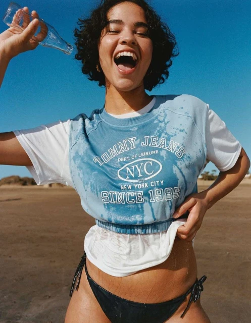Attractive female model in a Tommy Jeans shirt, wearing a bikini, holding a water bottle, outdoors on a beach on a sunny day.