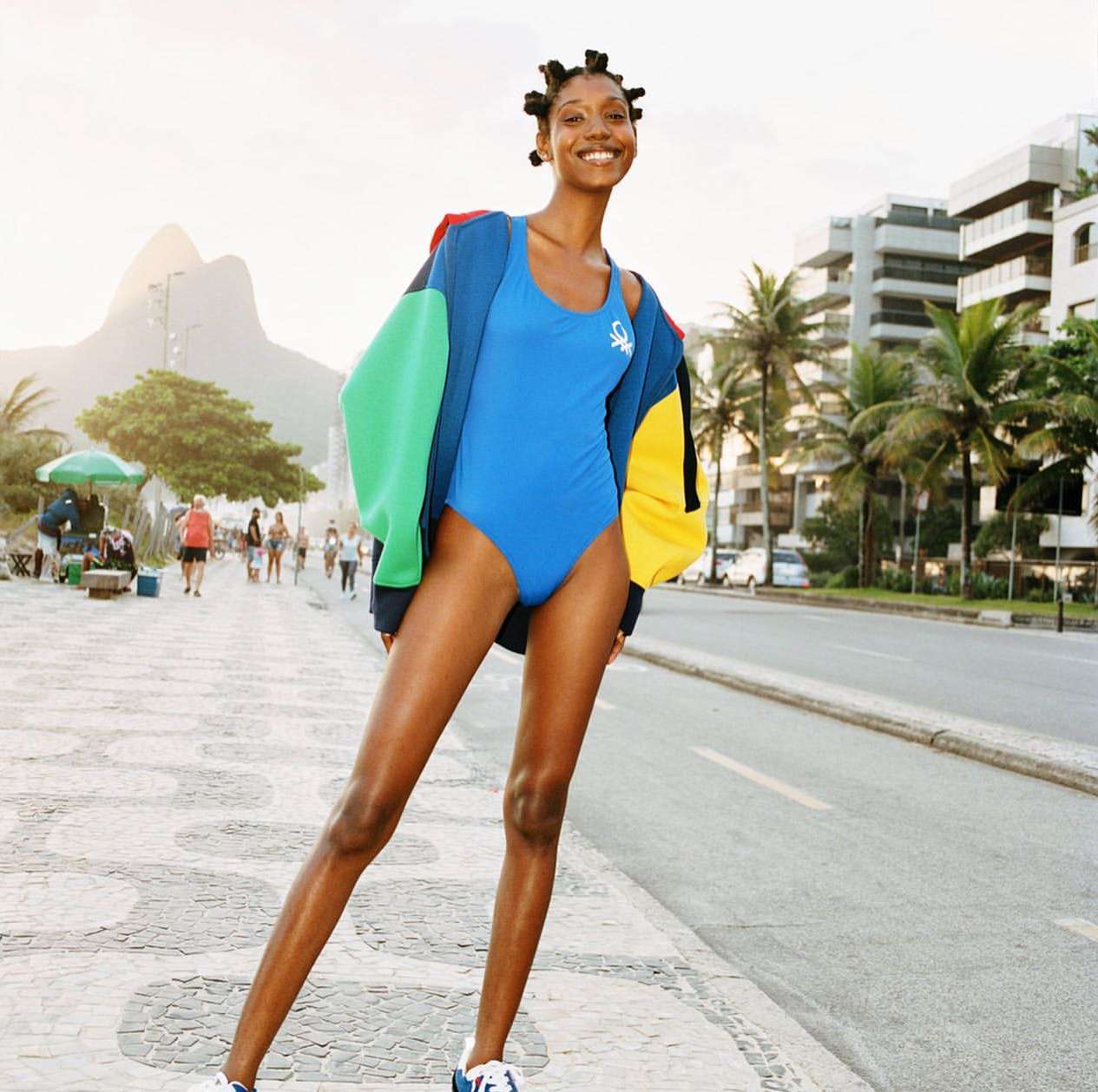 A woman in a blue swimsuit and color-block jacket stands on a patterned sidewalk in Rio de Janeiro, Brazil, smiling in a fashion editorial.