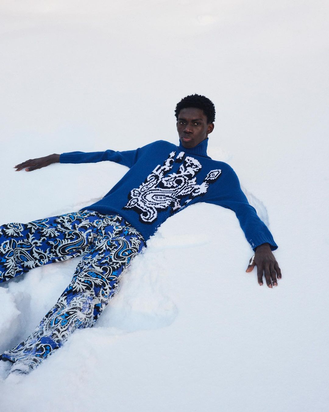Fashion shot of a Black male model in a blue patterned sweater and pants laying in the snow, creating a striking winter scene.