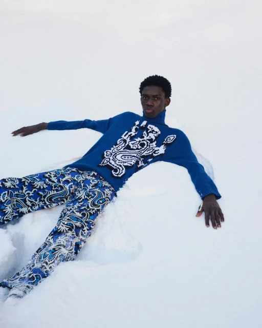 Fashion shot of a Black male model in a blue patterned sweater and pants laying in the snow, creating a striking winter scene.