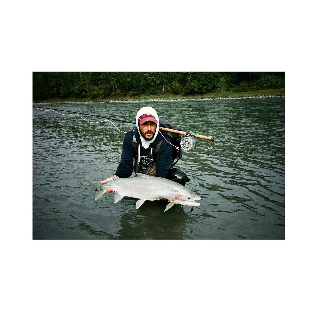 Man in river holding a large silvery salmon; he is wearing a hooded sweatshirt with a fly rod and reel setup over his shoulder.