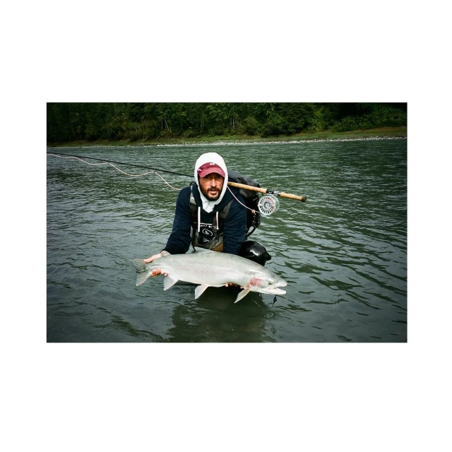Man in river holding a large silvery salmon; he is wearing a hooded sweatshirt with a fly rod and reel setup over his shoulder.