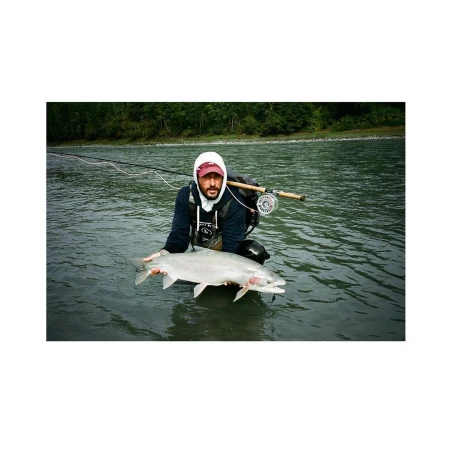 Man in river holding a large silvery salmon; he is wearing a hooded sweatshirt with a fly rod and reel setup over his shoulder.