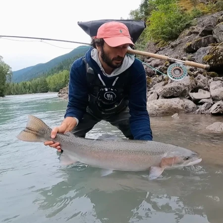 Angler holding a large trout in a clear river with a background of mountains and trees, showcasing outdoor fishing and nature.