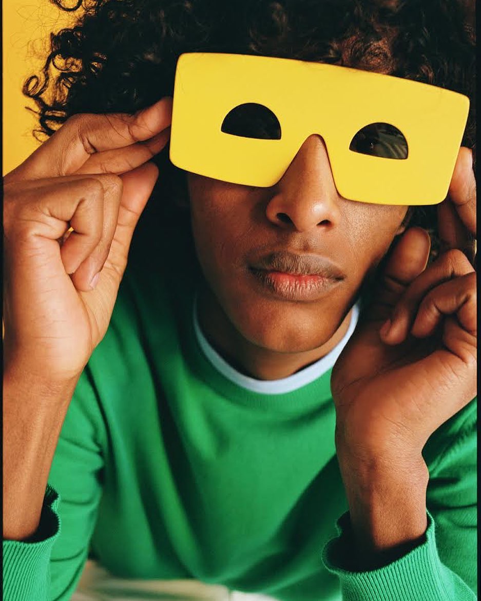 Close-up of an androgynous person with curly hair wearing a green sweater and yellow, rectangular novelty sunglasses against a yellow backdrop.