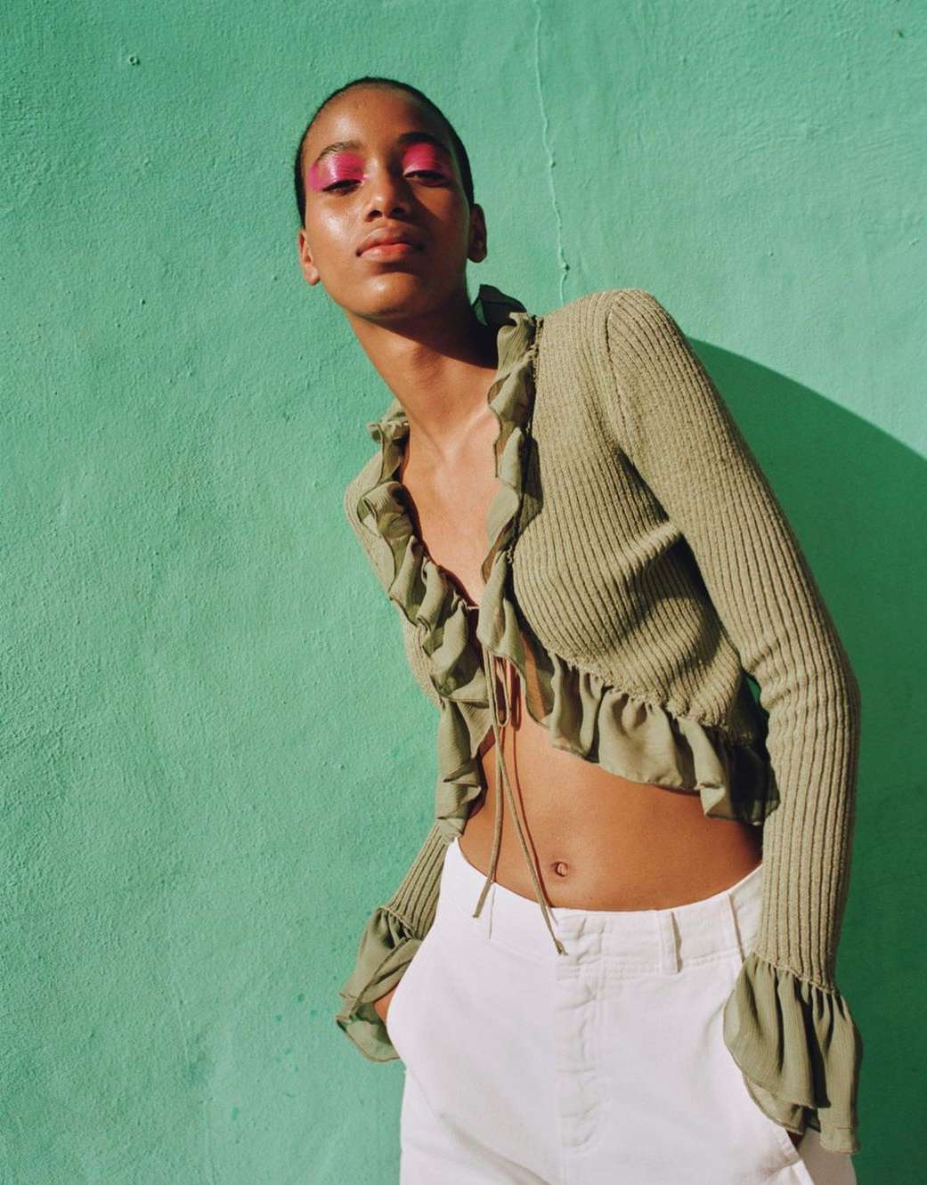 Model wearing a ruffled olive green cardigan and white trousers against a bright turquoise wall.