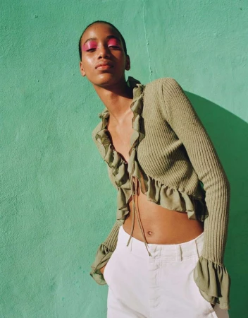 Model wearing a ruffled olive green cardigan and white trousers against a bright turquoise wall.