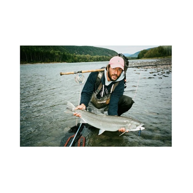 Fisherman poses with a large salmon in a river, surrounded by trees and hills under daylight. He wears a cap, waders, and fishing gear.