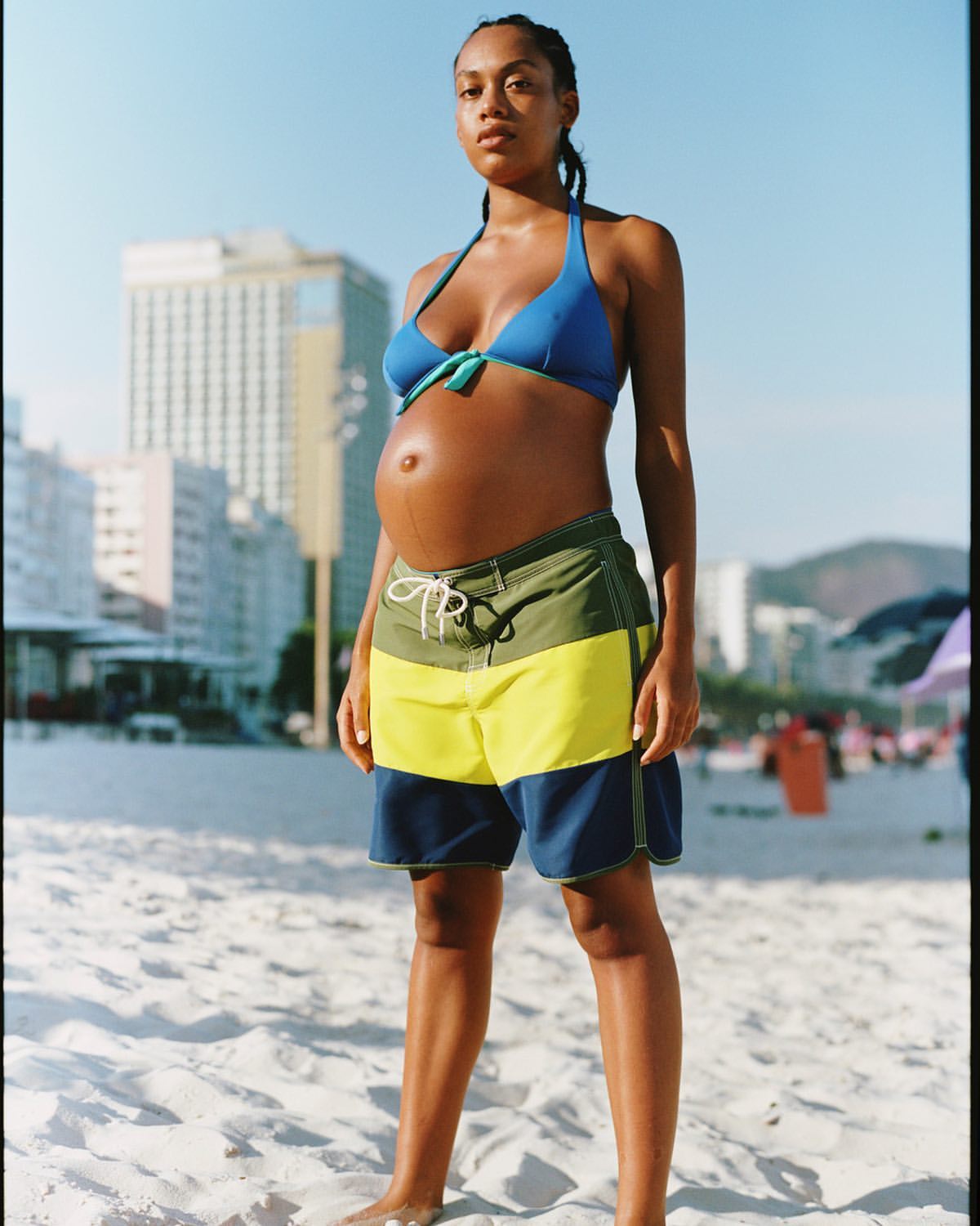 Pregnant woman wearing a blue bikini top and colour-blocked board shorts standing on a sandy beach under bright daylight.