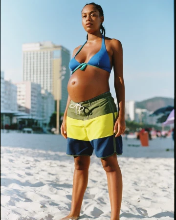 Pregnant woman wearing a blue bikini top and colour-blocked board shorts standing on a sandy beach under bright daylight.