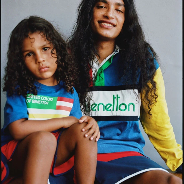 Two young fashion models pose indoors wearing Benetton shirts and shorts in a studio shot. They both have dark curly hair and dark complexions.
