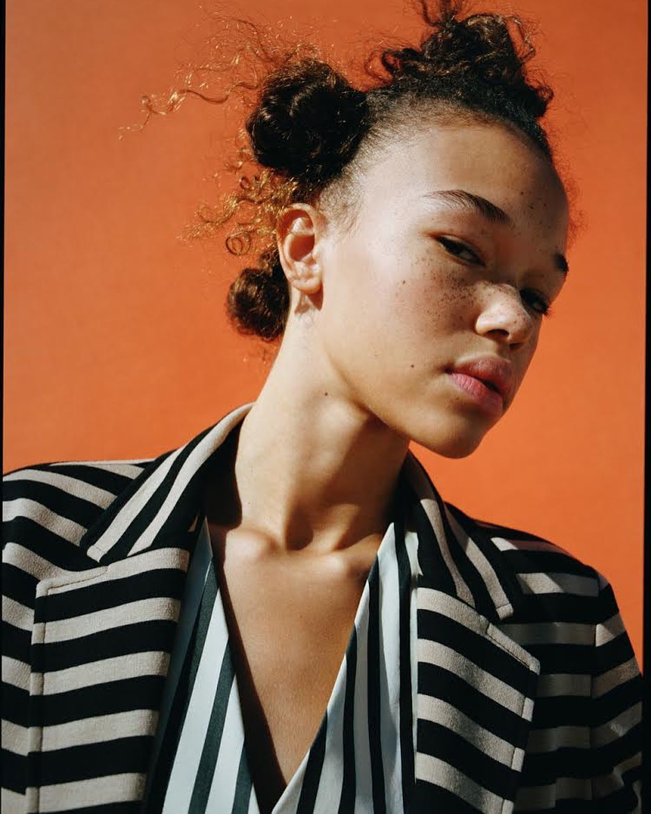 Close-up portrait of a young woman with freckles, wearing a striped blazer, set against a vibrant orange background.