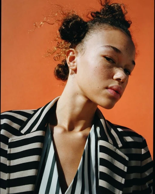 Close-up portrait of a young woman with freckles, wearing a striped blazer, set against a vibrant orange background.
