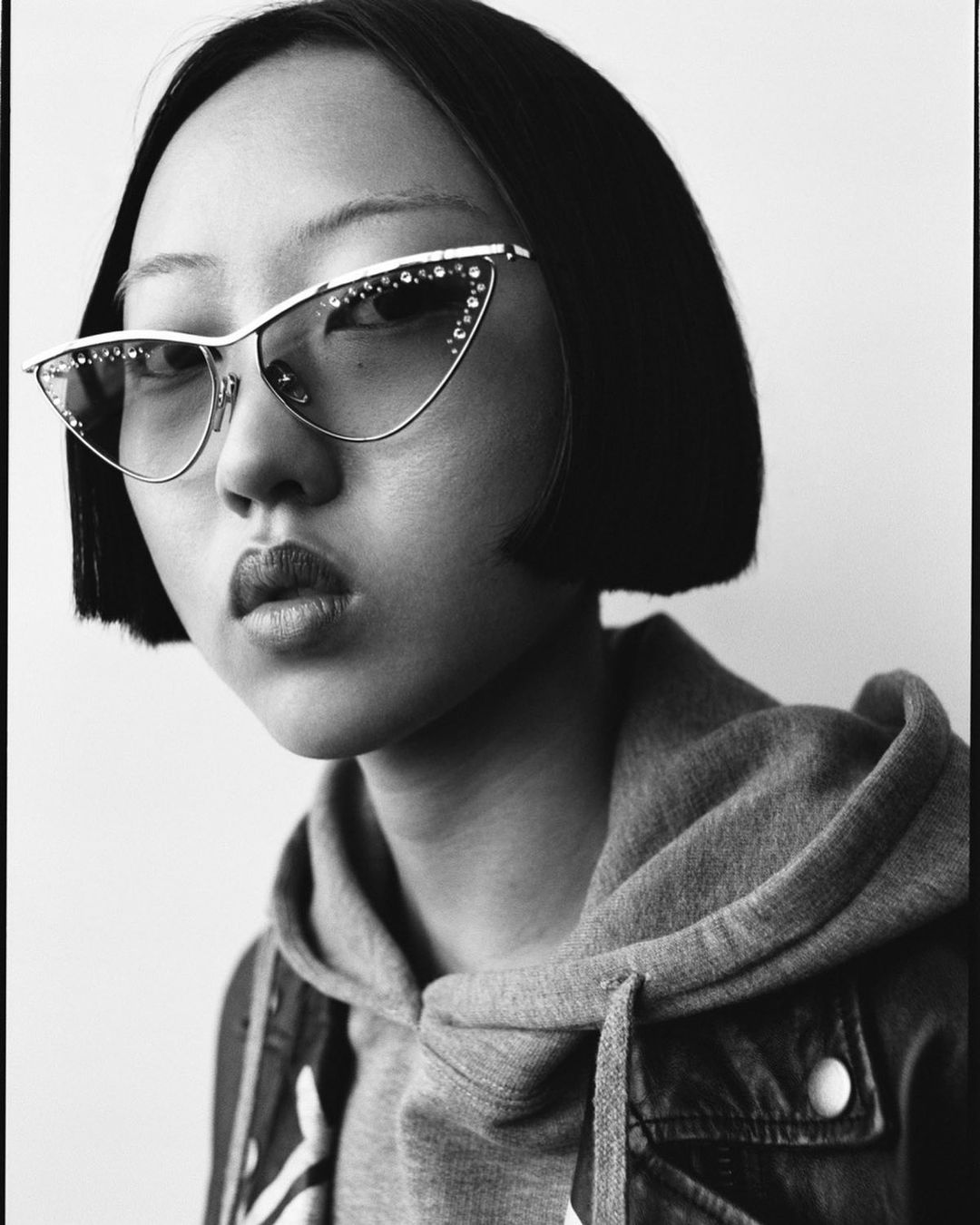 Black and white portrait of a young Asian woman wearing rhinestone cat eye glasses, hoodie and jean jacket in a simple studio setting.
