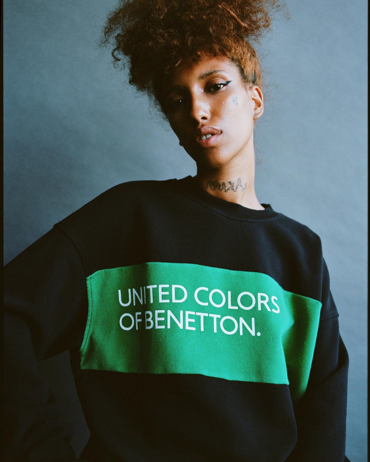 Woman in black and green United Colors of Benetton sweatshirt poses indoors against neutral grey background in studio lighting.