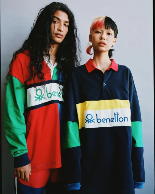 Two fashion models pose indoors wearing colorful long-sleeved polo shirts with the Benetton logo against a neutral gray background.