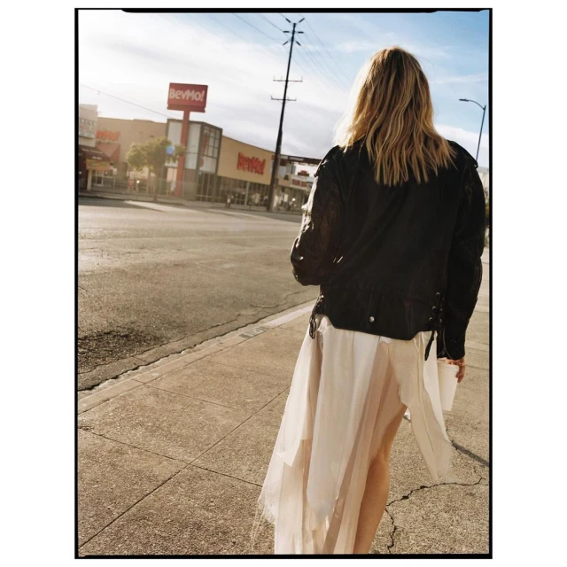 Woman in a black leather jacket and white dress walks on a city sidewalk on a sunny day. Buildings visible in the background.