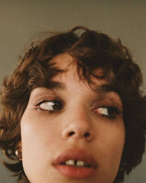 Close-up portrait of a young adult woman with curly brown hair, brown eyes, and light skin, looking slightly to the side in a studio setting.
