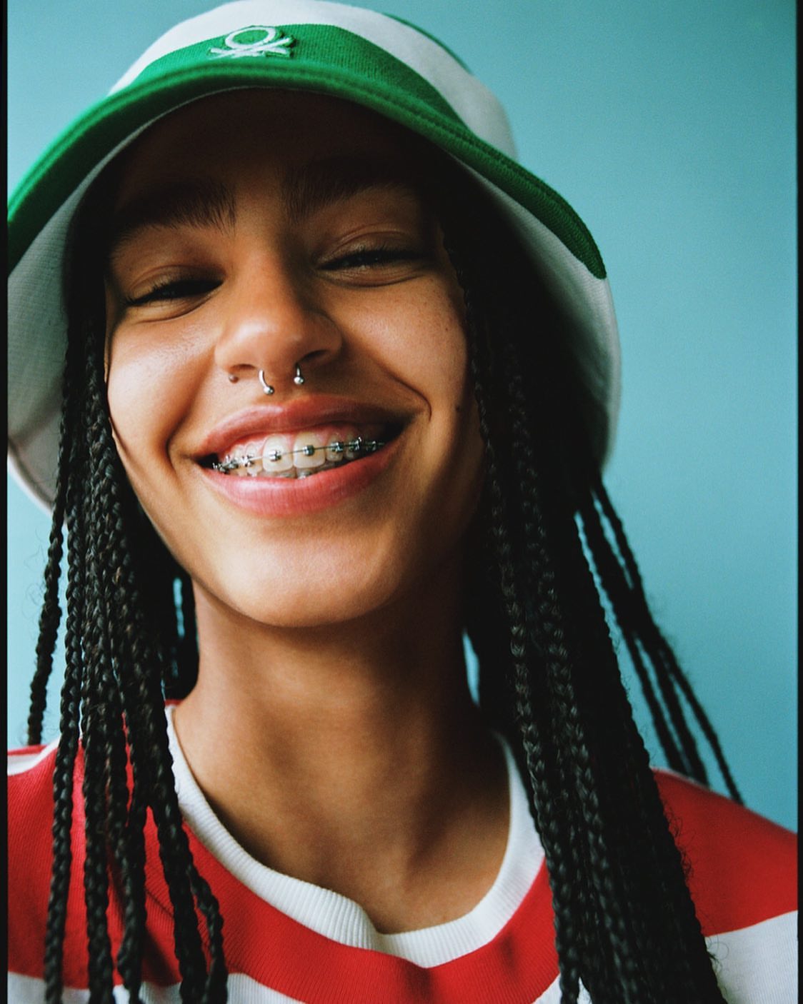 Close-up portrait of a smiling young woman with dark braids, braces, and a nose ring, wearing a green and white hat against a blue background.