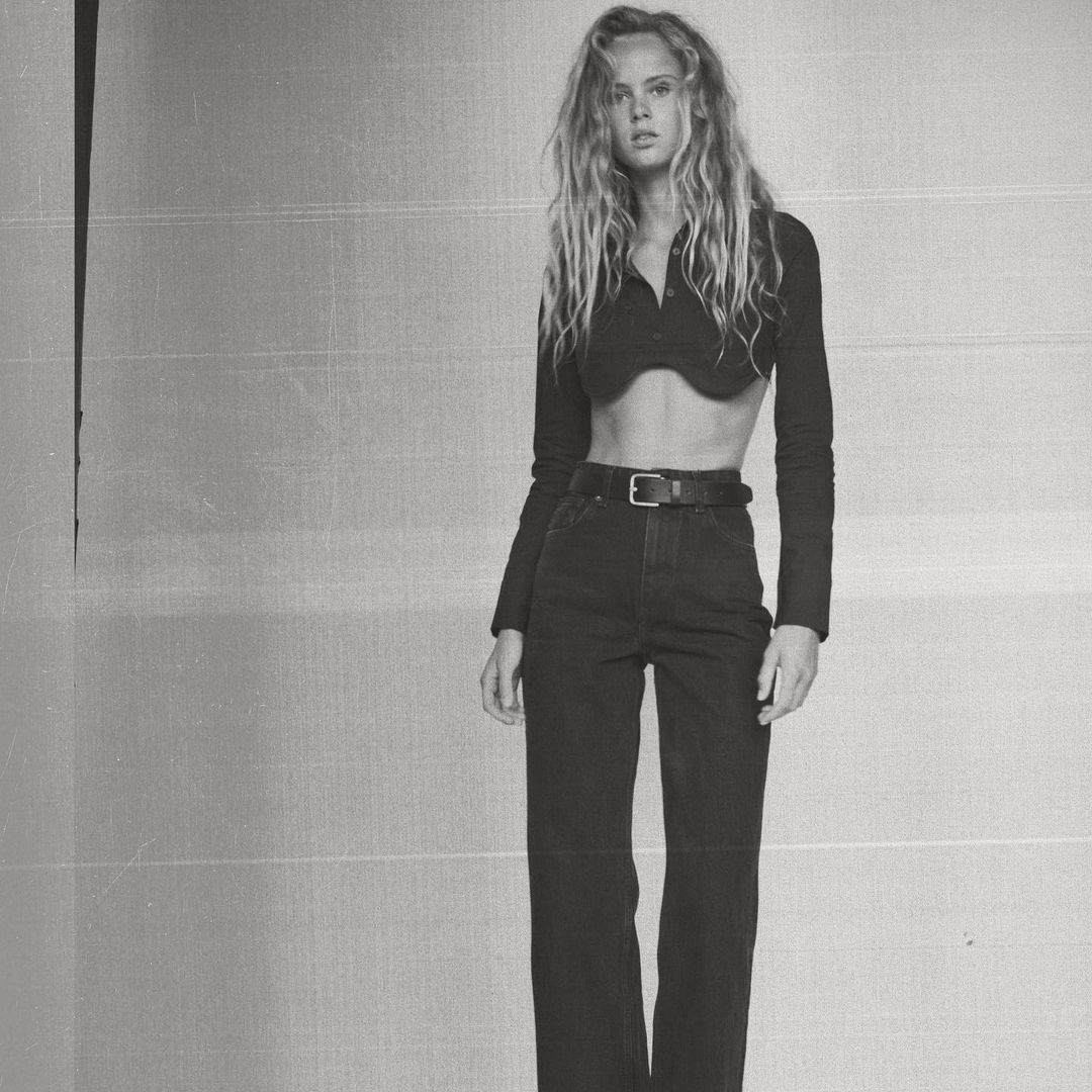 A young adult woman with wavy blonde hair poses in a cropped long sleeve shirt and dark jeans against a monochromatic textured background.