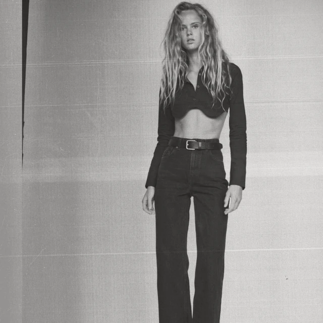A young adult woman with wavy blonde hair poses in a cropped long sleeve shirt and dark jeans against a monochromatic textured background.