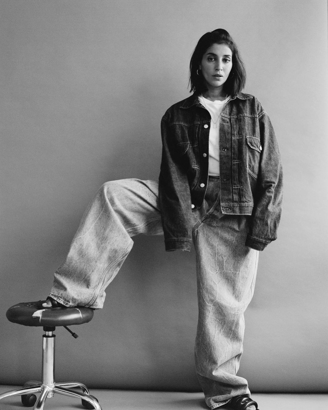 Woman in oversized denim jacket and jeans poses in a monochrome studio with a stool, creating a relaxed yet fashionable silhouette.