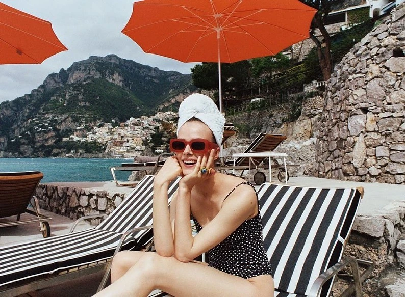 Woman in sunglasses and towel on head relaxing on a striped lounge chair by the coast with mountains and buildings in the background.