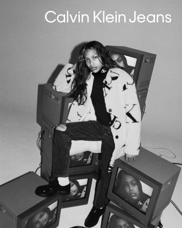 A young adult woman models for Calvin Klein Jeans, sitting on a stack of old television sets in a black and white fashion photograph.