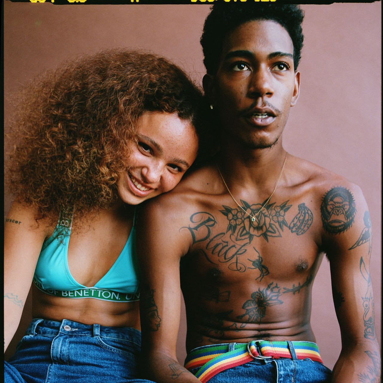 A young couple poses in a medium shot. The woman with curly hair rests her head on the man's shoulder, both wearing jeans.