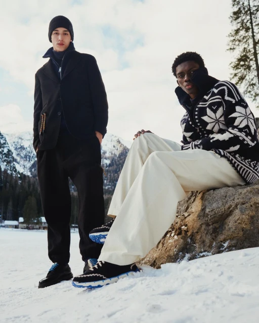 Two men showcase winter fashion in a snowy mountain setting, one seated on a rock in wide-leg pants and a patterned sweater, the other standing.
