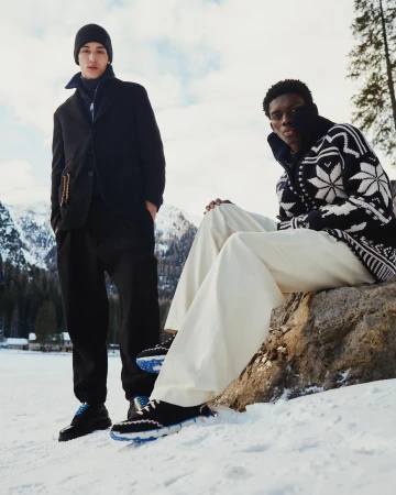Two men showcase winter fashion in a snowy mountain setting, one seated on a rock in wide-leg pants and a patterned sweater, the other standing.