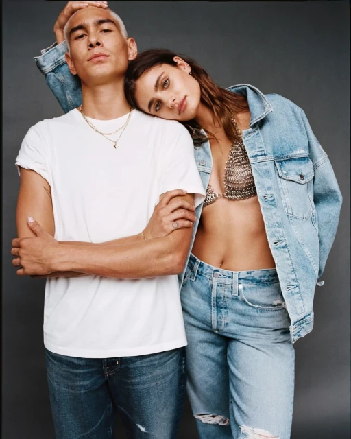 Two fashion models pose in light-wash denim, with the female model resting her head on the male model's head against a grey backdrop.