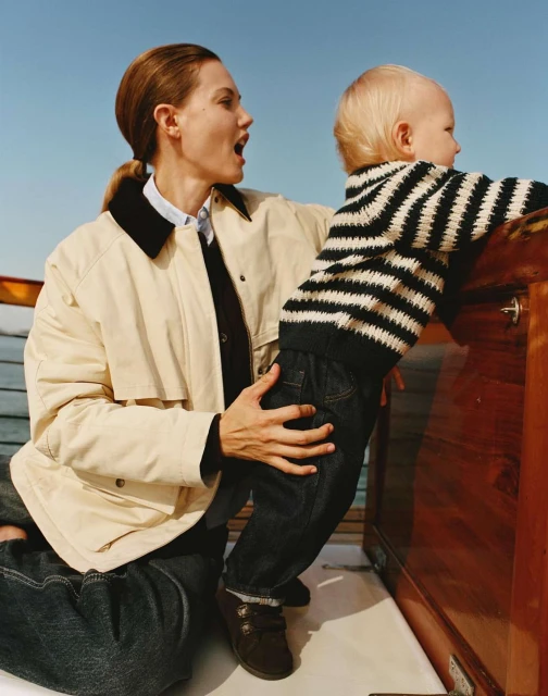 Mother and child on boat. Woman in tan jacket holds blonde child wearing striped sweater, standing near wood boat railing against sky.
