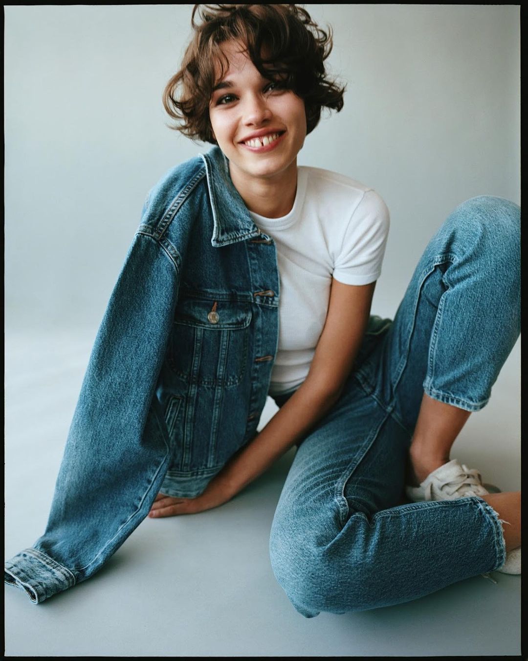 A smiling young woman in jeans and a white t-shirt is sitting on a grey surface with a blue jean jacket draped on her side.