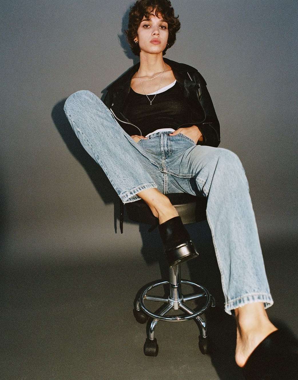 Fashionable young adult female poses indoors on chrome stool, wearing a black tank top, jacket, jeans, and slides against grey background.