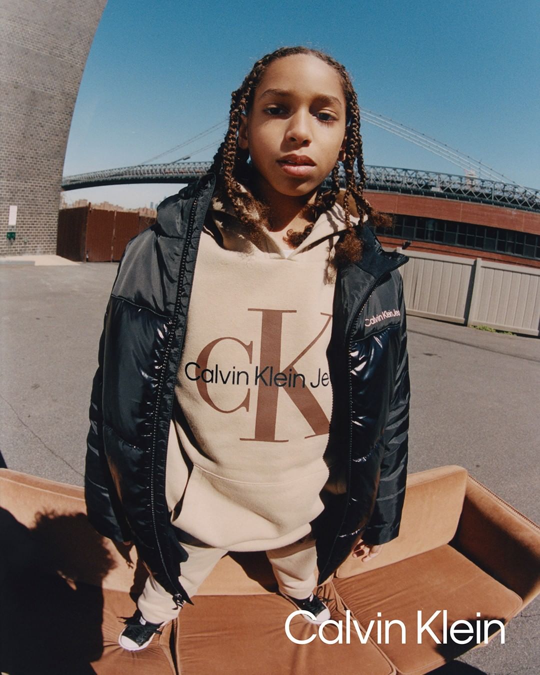 Person in Calvin Klein hoodie and puffer jacket poses on tan couch, with city bridge and blue sky backdrop during daytime.
