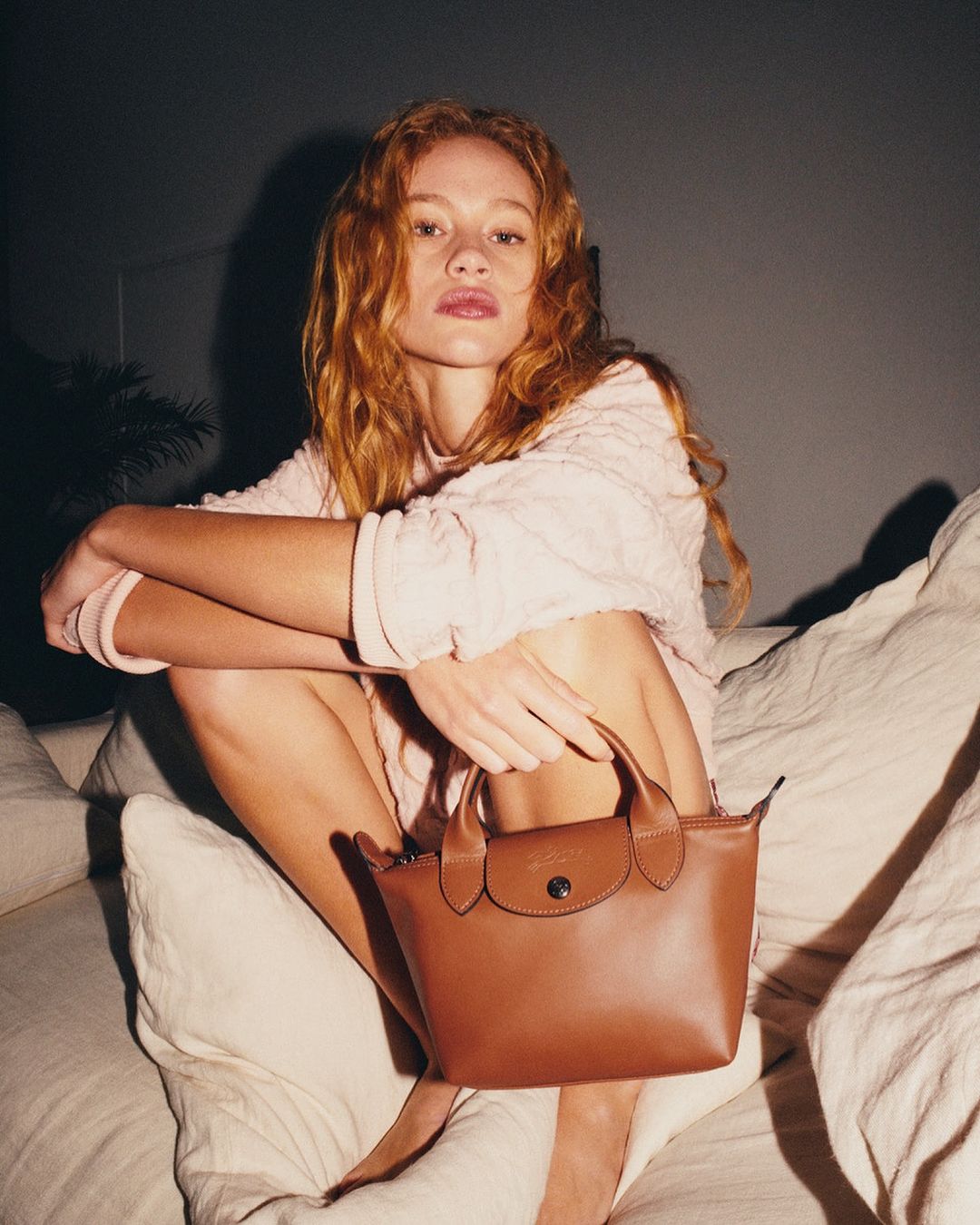 Red-haired woman in a light pink knit sweater holding a small brown leather handbag while seated on a white sofa.