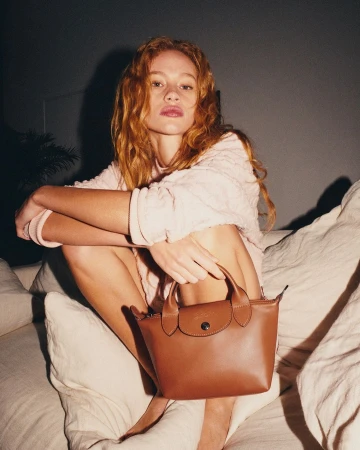 Red-haired woman in a light pink knit sweater holding a small brown leather handbag while seated on a white sofa.